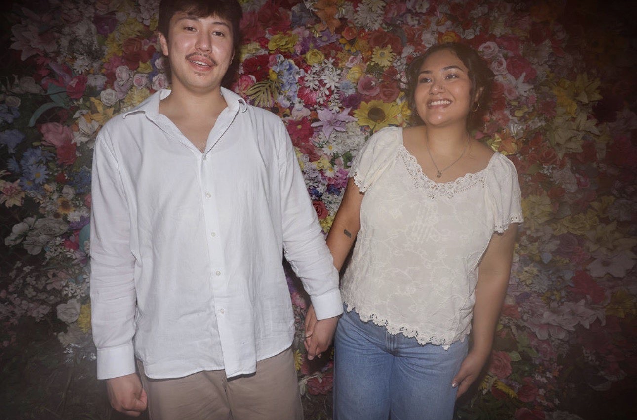 Ethan and Elli smiling in front of a wall of flowers